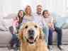 Ein Hund sitzt vor dem Sofa, auf dem seine Familie lächelnd nebeneinander sitzt.